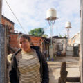 Josefina Tamara Villalba posa para una fotografía en la puerta de su casa en el barrio popular El Tambo. En el fondo, se ven los tanques de agua instalados con financiación del Fondo de Integración Socio Urbana. (Lucila Pellettieri/Global Press Journal)