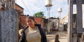 Josefina Tamara Villalba posa para una fotografía en la puerta de su casa en el barrio popular El Tambo. En el fondo, se ven los tanques de agua instalados con financiación del Fondo de Integración Socio Urbana. (Lucila Pellettieri/Global Press Journal)